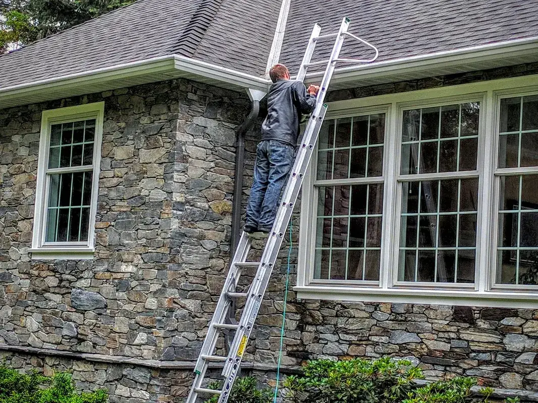 A worker on an extension ladder cleaning the gutter of a stone house